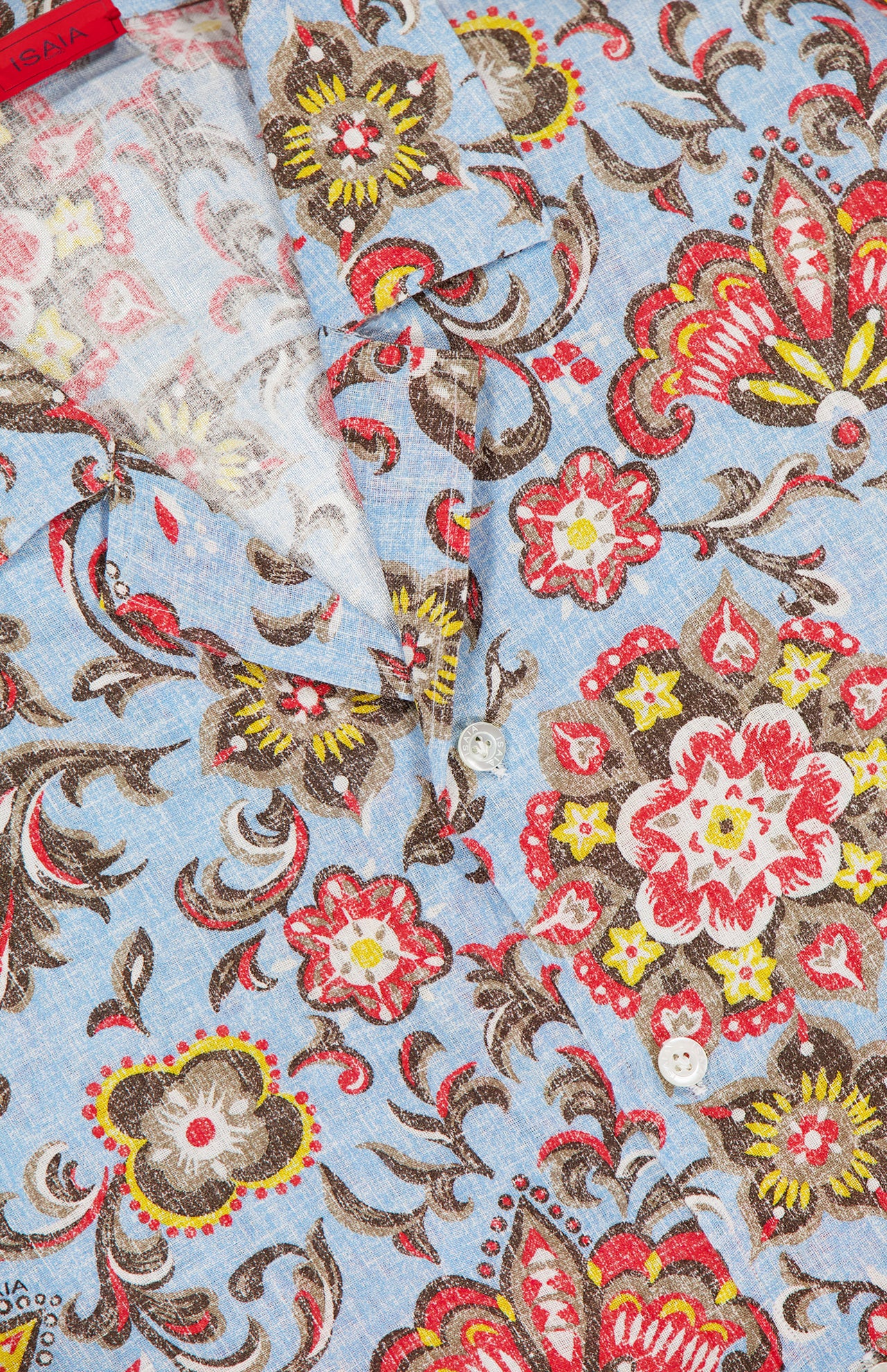 Close-up of the ISAIA Superlino Print Linen Floral Camp Shirt in blue, featuring a multicolor paisley pattern, white buttons partially unfastened, and a visible red brand tag inside the collar.