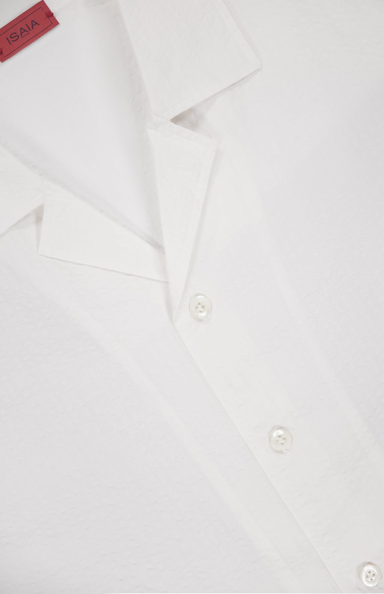 Close-up of the ISAIA Seersucker Short Sleeve Camp Shirt featuring a textured white finish, spread collar, and three white buttons. A red ISAIA tag is partially visible at the top left.