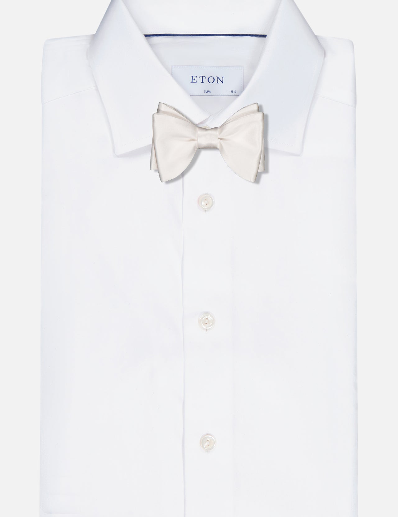A neatly folded white dress shirt with an ITALO FERRETTI Satin Bowtie in light cream, handmade in Italy, displayed against a plain white background. The shirt has front buttons and a classic pointed collar.