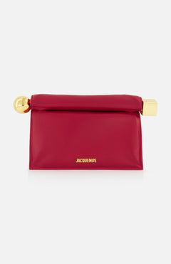 The JACQUEMUS La Pochette Rond Carre Clutch is a rectangular, deep pink French-style bag featuring a fold-over top, gold hardware at both ends, and JACQUEMUS in gold on the front—a bold designer statement for any event.