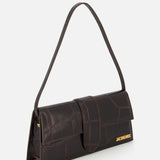 The JACQUEMUS Le Bambino Long Croco-effect Shoulder Bag features dark brown leather, a rectangular shape, flap closure, single shoulder strap, and gold logo at the bottom right corner, shown against a plain white background.