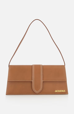 The Le Bambino Long Shoulder Bag by JACQUEMUS is a brown rectangular leather bag with a top handle, single shoulder strap, white stitching, front flap closure, and gold Jacquemus logo at the bottom right. Made in Italy.