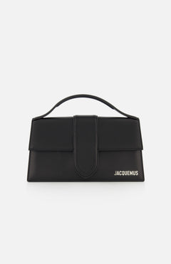 The Le Grand Bambino Bag by JACQUEMUS is a black leather top-handle bag with a flap closure, adjustable crossbody strap, and the JACQUEMUS logo in white on the lower right—made in Italy and shown against a white background.