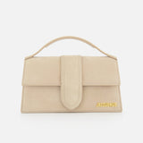 The JACQUEMUS Le Grand Bambino Bag is a tan leather top-handle bag made in Italy, featuring a front flap closure and gold logo at the bottom right corner, shown against a white background.