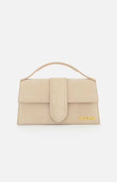 The JACQUEMUS Le Grand Bambino Bag is a tan leather top-handle bag made in Italy, featuring a front flap closure and gold logo at the bottom right corner, shown against a white background.