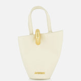 The Le Petit Bambola Tote Bag by JACQUEMUS is a small, structured off-white handbag with dual handles, gold hardware, and “JACQUEMUS” in gold lettering on the front for a minimalist, elegant look.