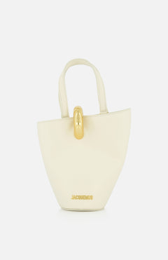The Le Petit Bambola Tote Bag by JACQUEMUS is a small, structured off-white handbag with dual handles, gold hardware, and “JACQUEMUS” in gold lettering on the front for a minimalist, elegant look.