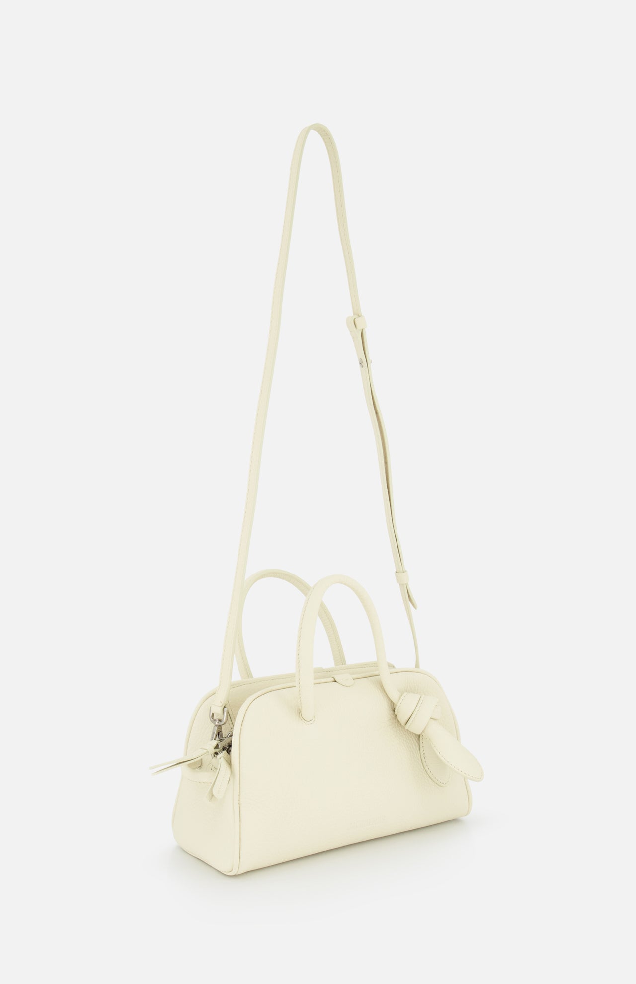 The JACQUEMUS Le Petit Turismo Bowling Bag is a small, off-white leather accessory with two handles, an adjustable strap, zipper closure, and a knotted detail—capturing quiet luxury against a plain white background.