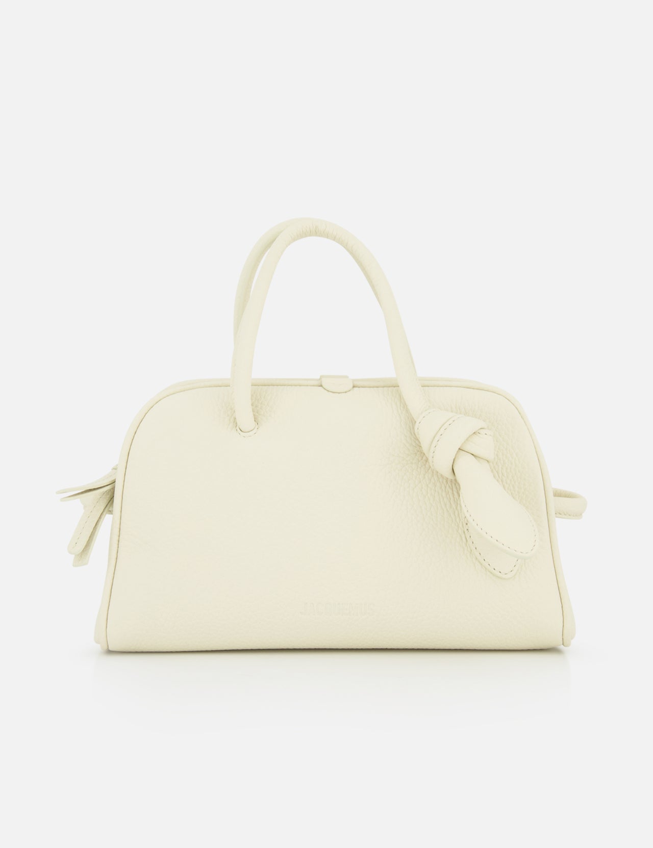 The Le Petit Turismo Bowling Bag by JACQUEMUS is a small, off-white leather accessory with two top handles and a side knotted strap, embodying quiet luxury against a plain white backdrop.