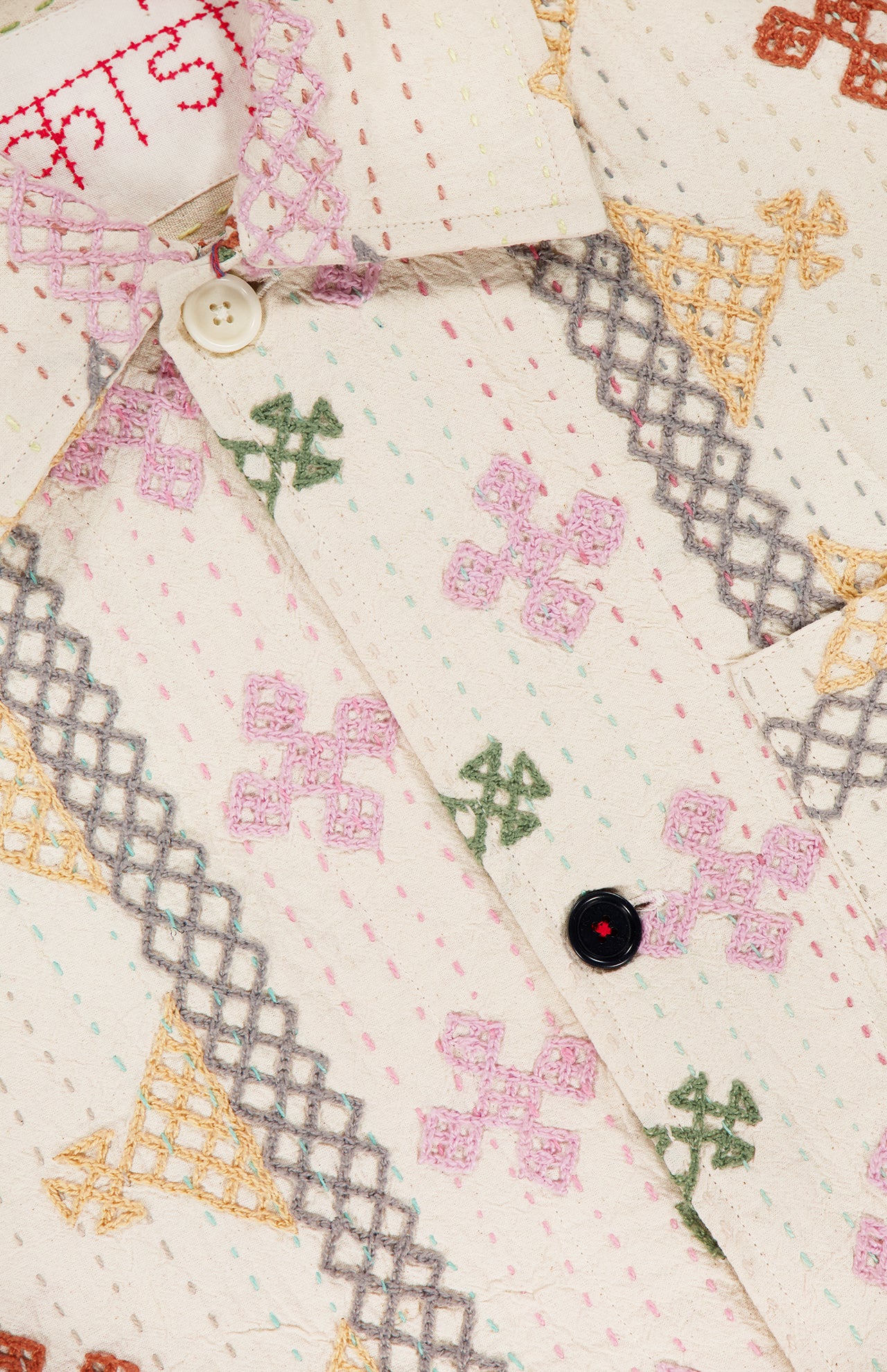 Close-up of the KARDO Bodhi Shirt Jacket, made from cream-colored 100% cotton with pink, green, yellow, and gray embroidered geometric patterns, visible stitching, two buttons, and a partially shown fabric label indicating it's made in India.