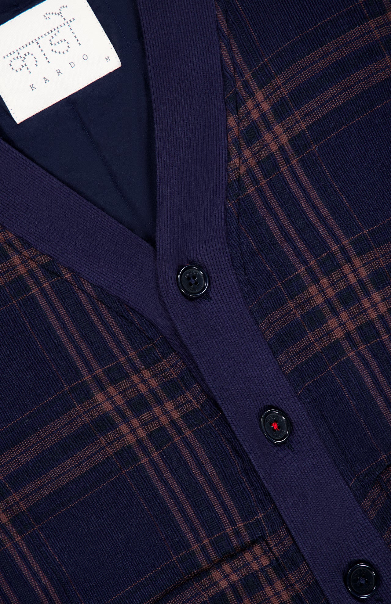 Close-up of the Janesh Quilted Plaid Cardigan by KARDO, featuring a dark blue and brown plaid pattern, black buttons, ribbed navy trim, and an interior white label with “KARDO M” and additional script. This men’s cardigan offers classic style blended with everyday comfort.