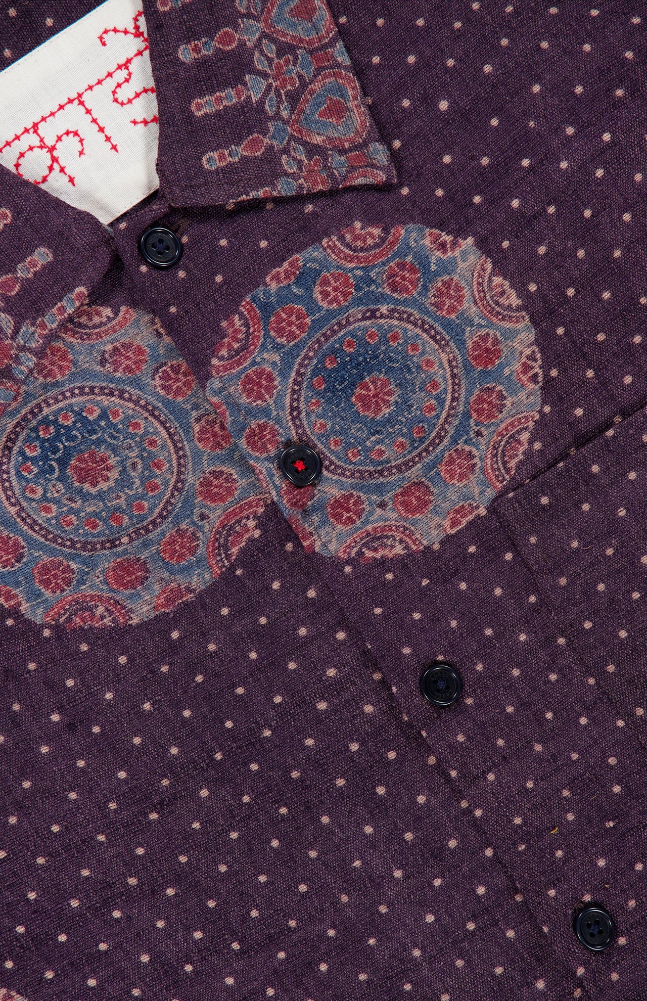 A close-up of the KARDO Martin Relaxed Fit Long Sleeve Shirt in purple, showcasing small white polka dots, dark buttons, and circular blue and red designs near the collar and chest. A portion of a white label with red stitching is also visible.