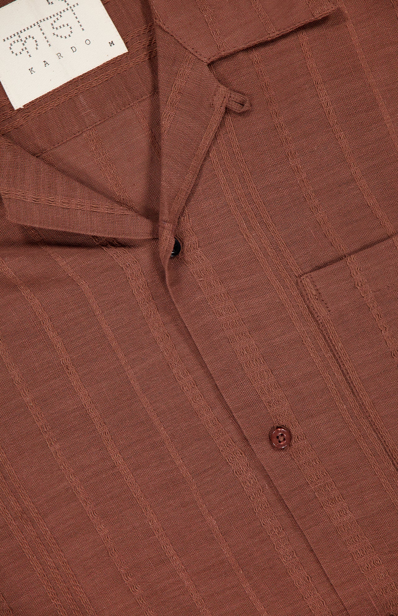 Close-up of the KARDO Rohit Stripe Short Sleeve Shirt in brown 100% cotton, featuring a subtle stripe pattern, oversized fit, chest pocket, visible buttons, and a partially visible white label at the collar.