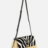 The Audrey Raffia Crossbody Bag by KHAITE features a handwoven straw base from Madagascar, a black and white animal print flap, a small gold front clasp, and a long adjustable black leather strap.