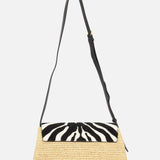 The KHAITE Audrey Raffia Crossbody Bag features handwoven Madagascar raffia, a black adjustable strap, and a flap with a striking black and white animal print motif.