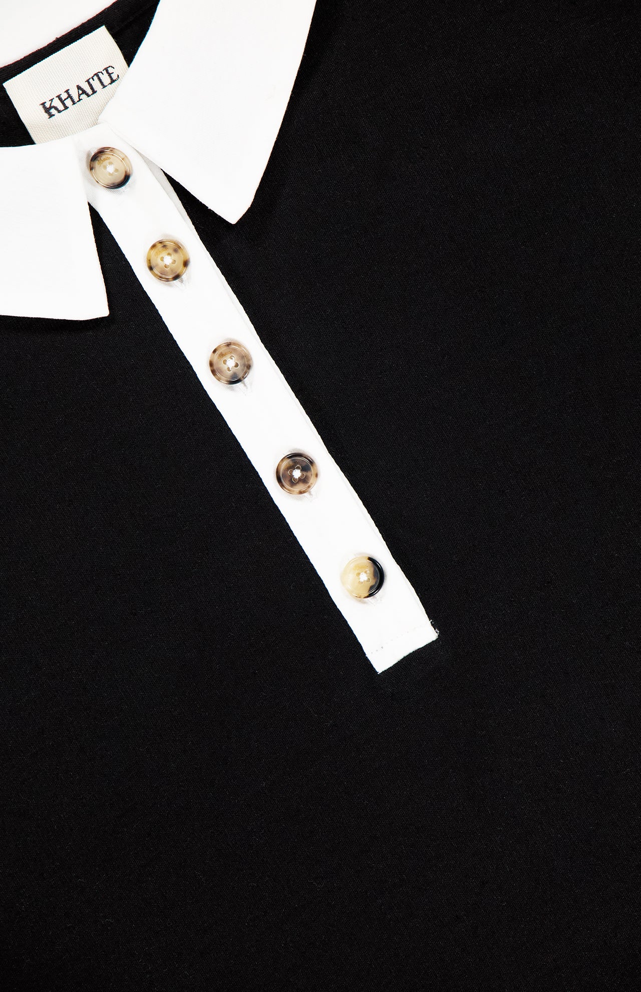 Close-up of the KHAITE Faithful Vintage Rugby Top, a black unisex silk polo shirt with a white collar and four marbled buttons on its white placket. The inner collar label displays "KHAITE.