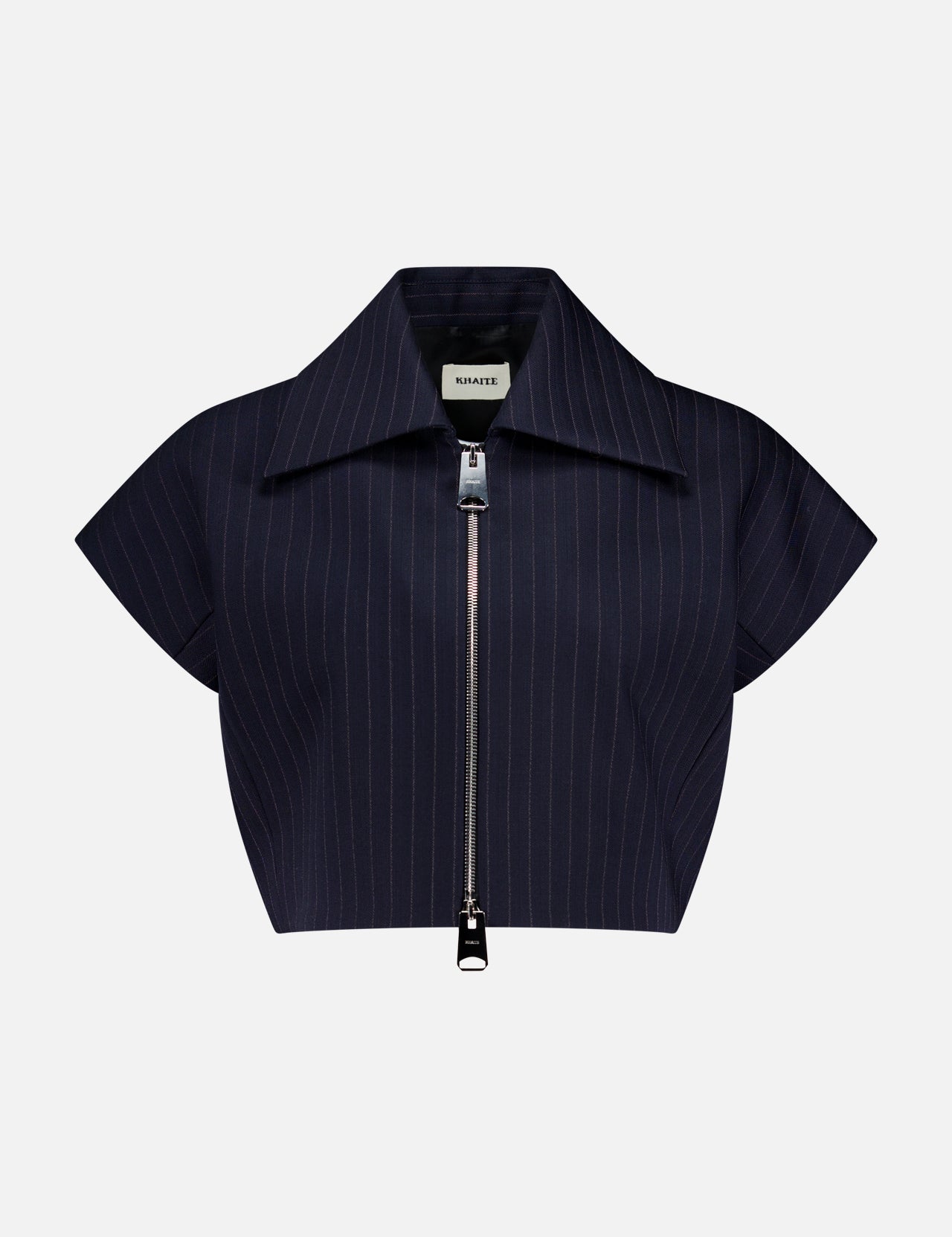 The Robin Striped Crop Top by KHAITE features navy and white stripes, short sleeves, a wide collar, and a front silver zipper in suiting fabric. The background is plain light gray.
