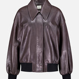 The Shelman Leather Bomber Jacket by KHAITE features a dark brown leather design with a wide collar, snap button closure, and black ribbed cuffs and hem, displayed against a plain white background.
