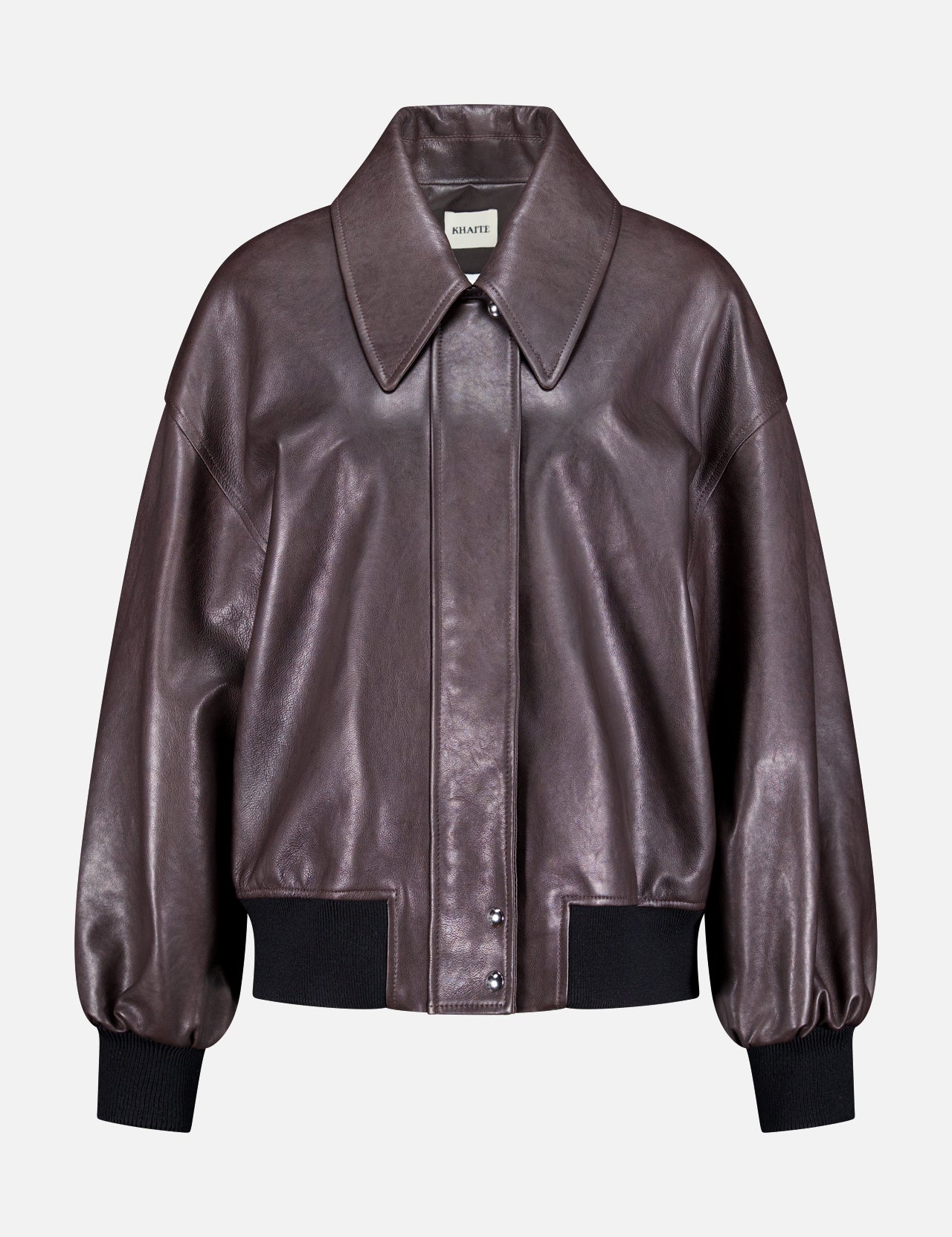The Shelman Leather Bomber Jacket by KHAITE features a dark brown leather design with a wide collar, snap button closure, and black ribbed cuffs and hem, displayed against a plain white background.
