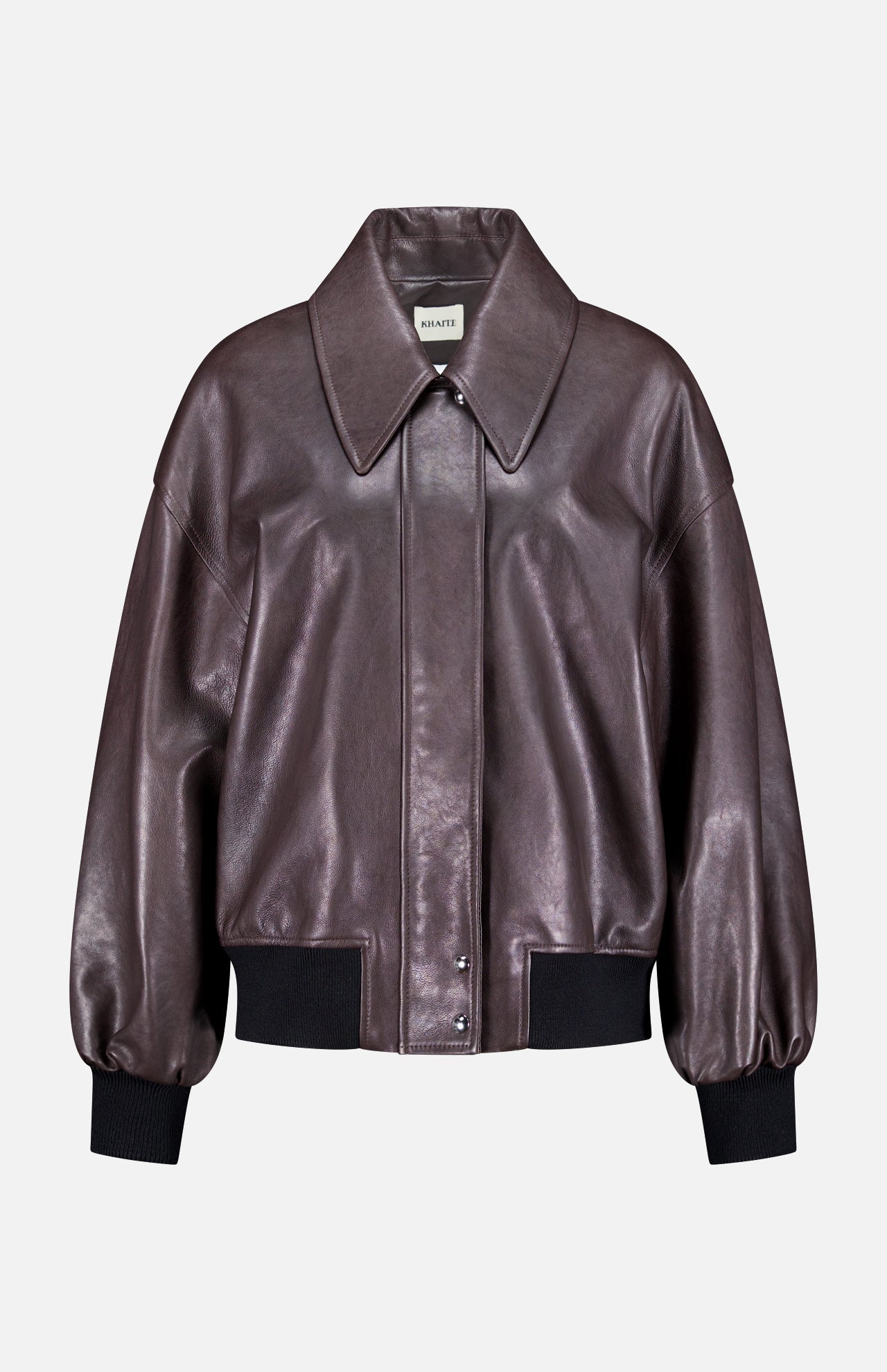 The Shelman Leather Bomber Jacket by KHAITE features a dark brown leather design with a wide collar, snap button closure, and black ribbed cuffs and hem, displayed against a plain white background.