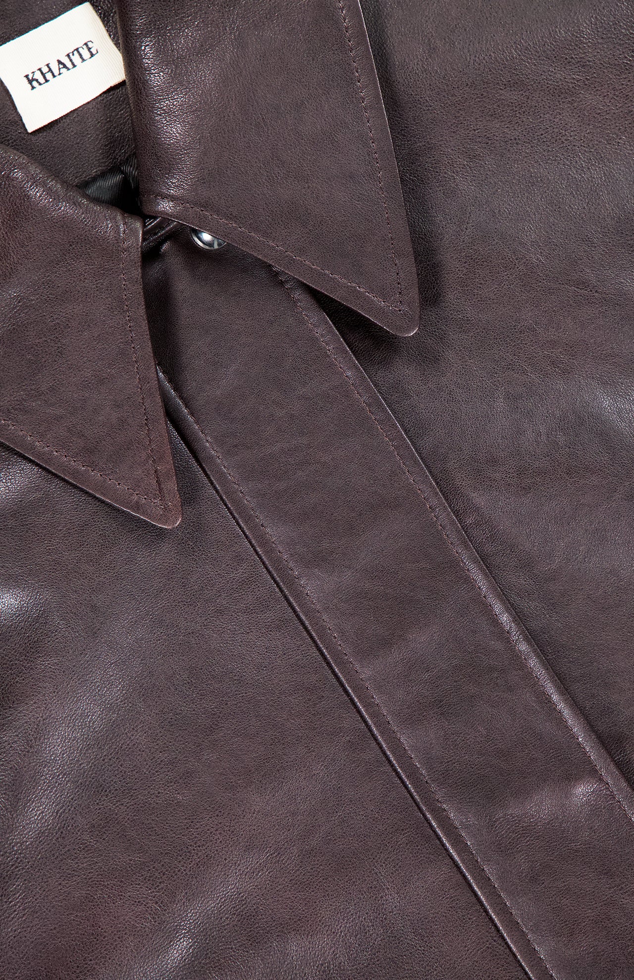 Close-up of the Shelman Leather Bomber Jacket by KHAITE, shown in dark brown lambskin with a pointed collar, visible stitching, and a label displaying the KHAITE brand name in the top left corner.