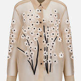 The Argo Top with Floral Embroidery by KHAITE is a sheer beige silk organza blouse featuring a collar, white floral appliqué, and black embroidered stems—an elegant women’s statement piece.