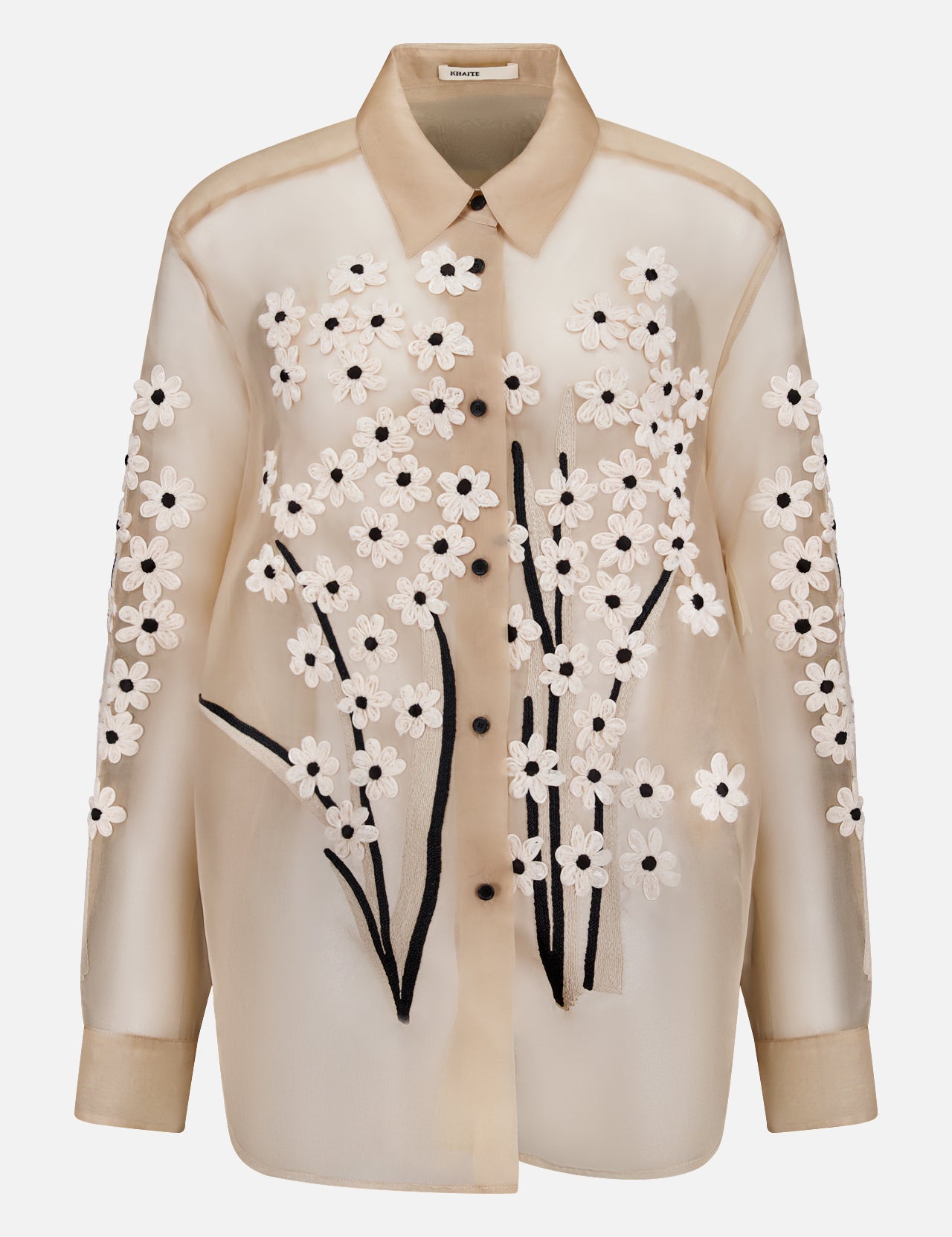 The Argo Top with Floral Embroidery by KHAITE is a sheer beige silk organza blouse featuring a collar, white floral appliqué, and black embroidered stems—an elegant women’s statement piece.