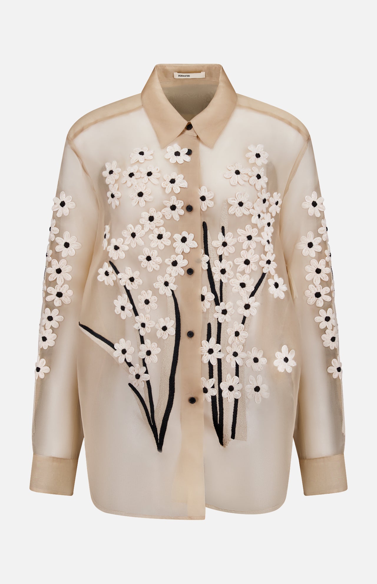 The Argo Top with Floral Embroidery by KHAITE is a sheer beige silk organza blouse featuring a collar, white floral appliqué, and black embroidered stems—an elegant women’s statement piece.