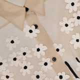 Close-up of the KHAITE Argo Top with Floral Embroidery, a sheer silk organza blouse featuring black buttons and embroidered white flowers with black centers. The visible collar and stitching accentuate this elegant women's statement piece.