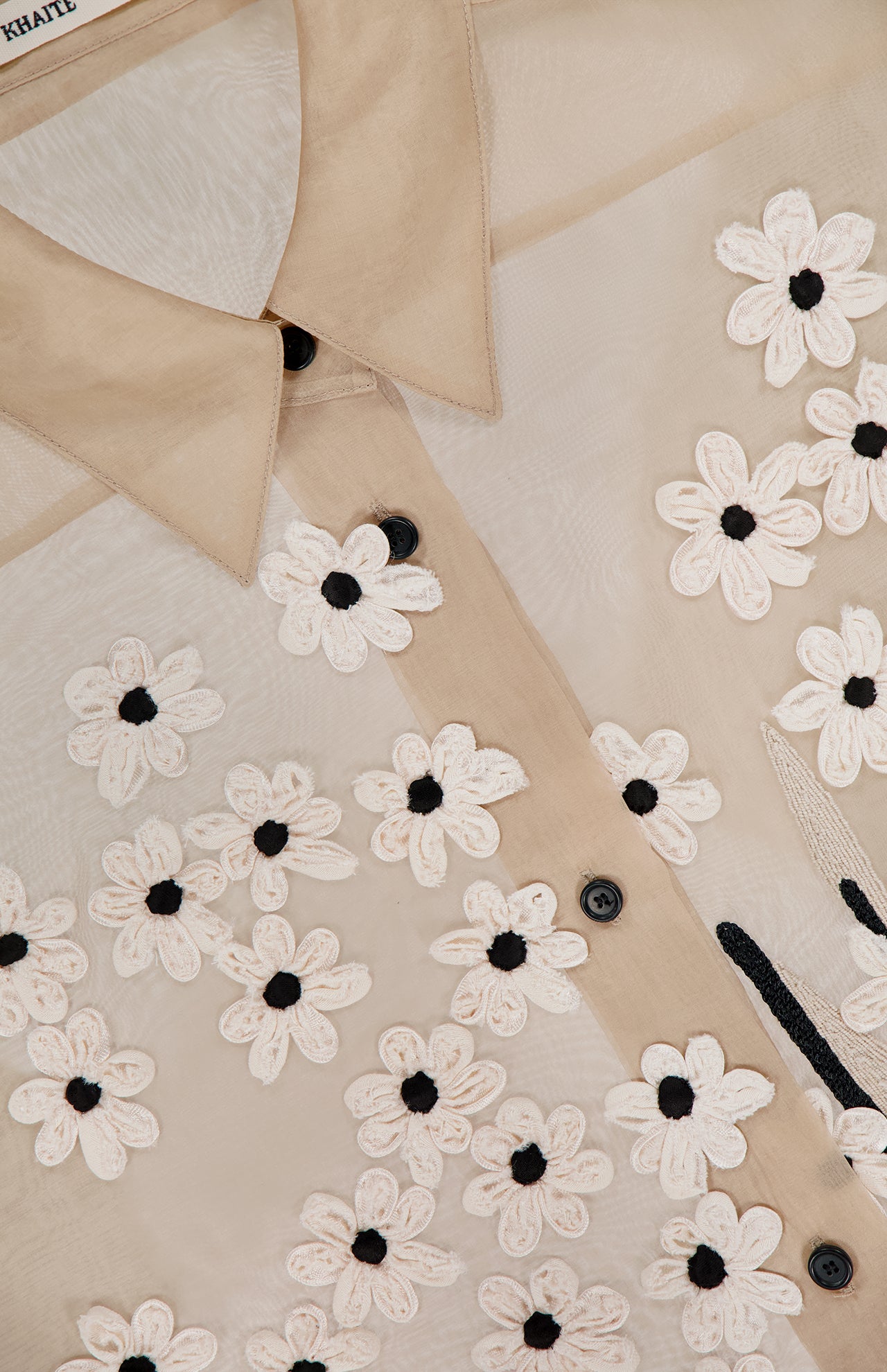 Close-up of the KHAITE Argo Top with Floral Embroidery, a sheer silk organza blouse featuring black buttons and embroidered white flowers with black centers. The visible collar and stitching accentuate this elegant women's statement piece.