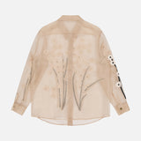 The KHAITE Argo Top with Floral Embroidery is a sheer beige silk organza blouse with a collar, buttoned cuffs, and delicate floral embroidery on the back and one sleeve—an elegant women’s statement piece crafted from lightweight, translucent fabric.