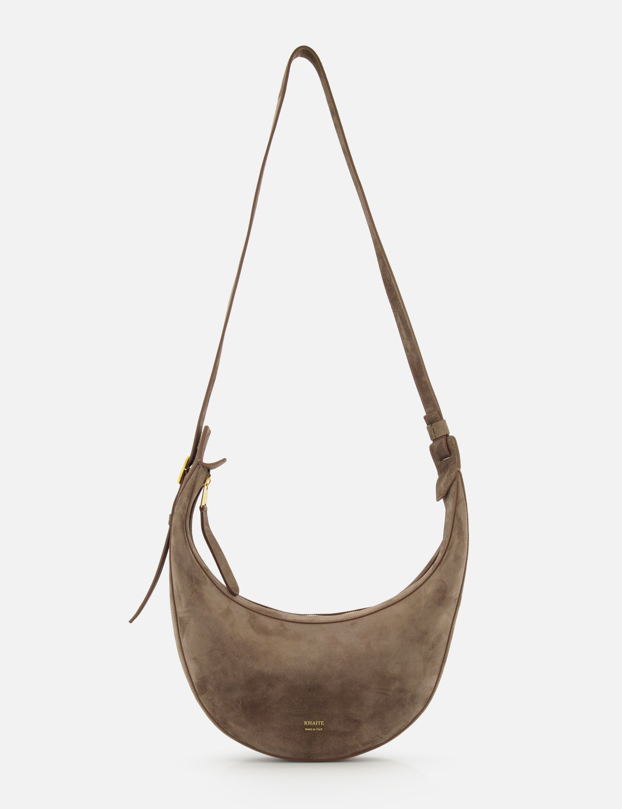 The Augustina Small Crossbody Bag by KHAITE is a light brown suede bag with a curved zip-top hobo silhouette, adjustable strap, gold hardware, and embossed logo on the front—crafted in Italy and shown on a white background.