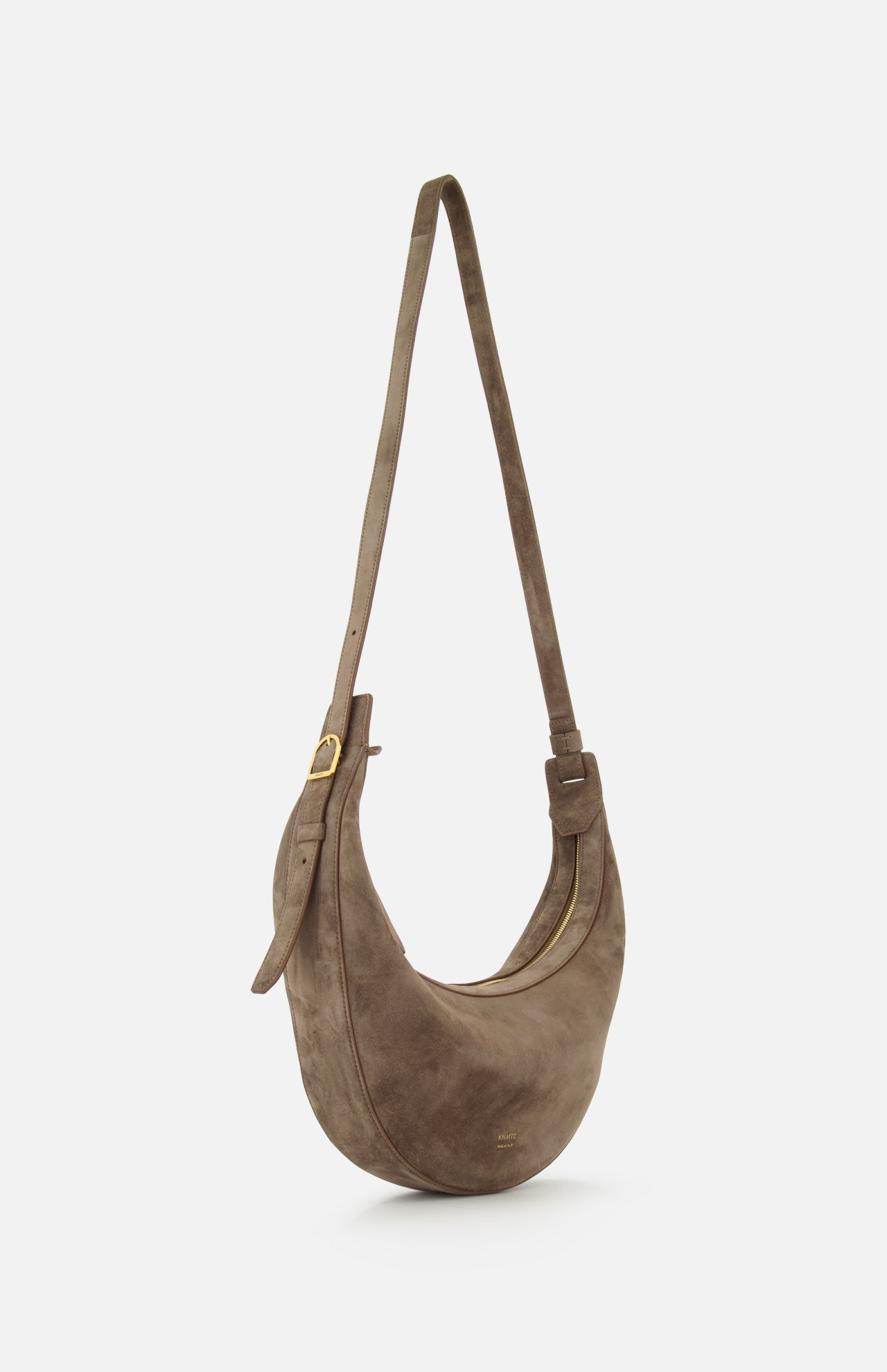 The Augustina Small Crossbody Bag by KHAITE features light brown suede in a crescent, zip-top hobo silhouette with a long adjustable strap and gold-tone hardware. Made in Italy, it's shown against a plain white background.