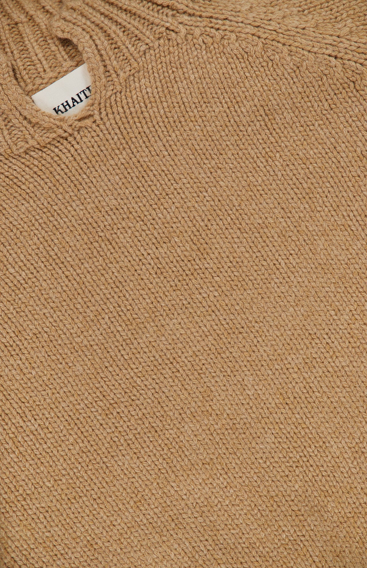 Close-up of the beige Cain Split Neck Cashmere Sweater by KHAITE, showcasing its textured stitching and a small white label near the neckline—a luxurious Italian cashmere choice for any minimalist wardrobe.