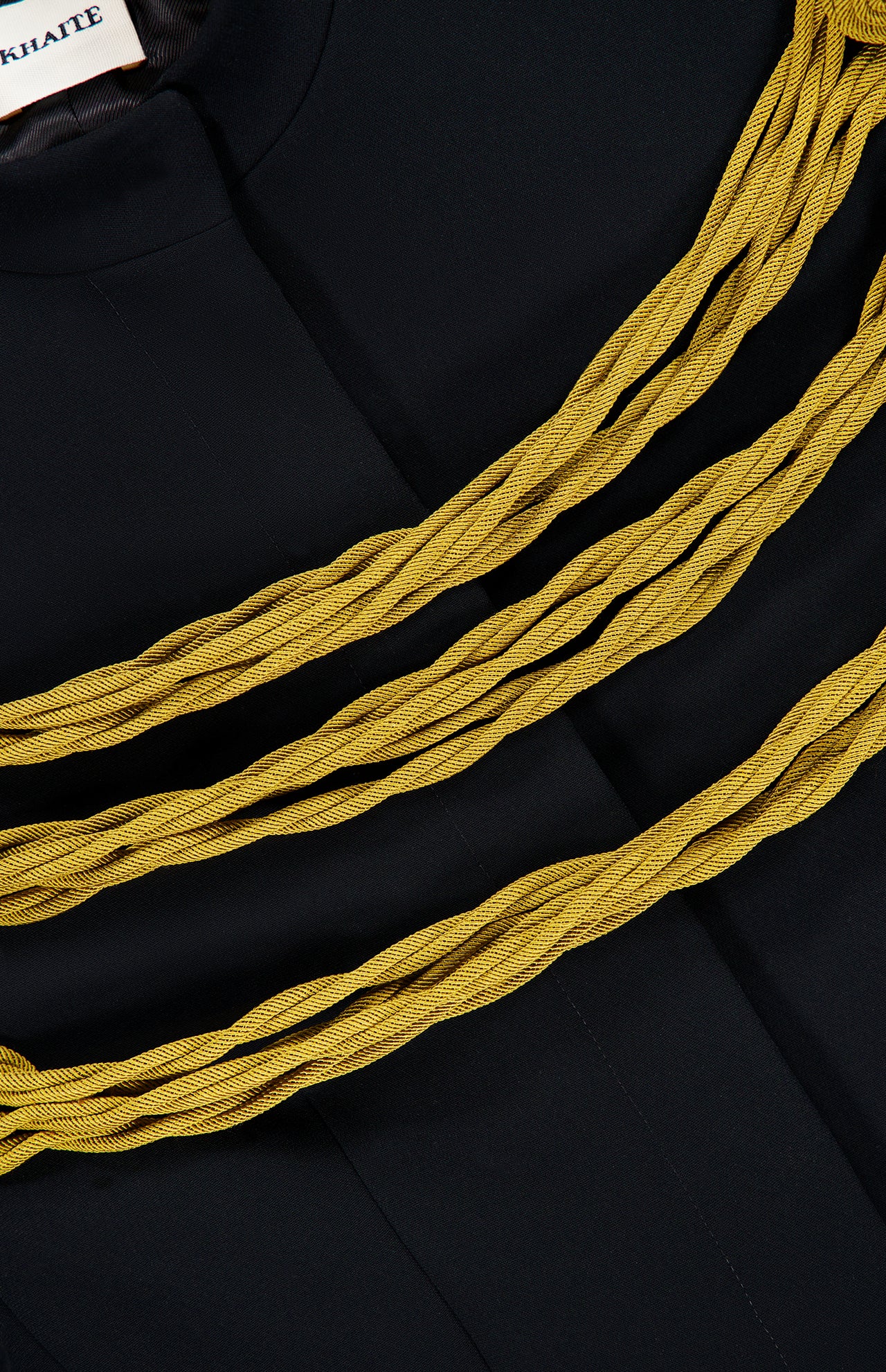 Close-up of the KHAITE Cord Embroidery York Jacket in black, featuring three thick, twisted yellow cords arranged across the chest in a style reminiscent of gold cord embroidery. A partially visible white KHAITE label with black text appears in the upper left corner.