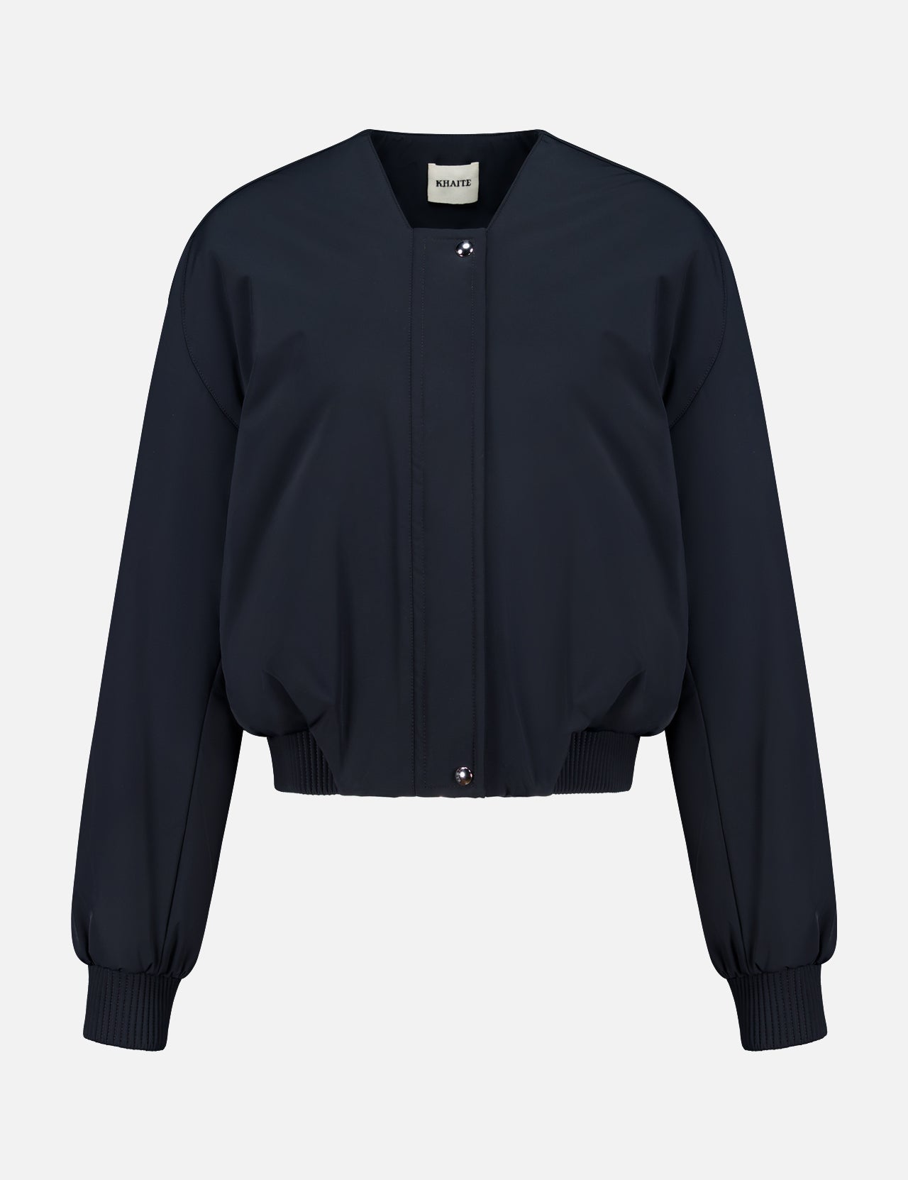 The Cordan Cropped Bomber Jacket by KHAITE is showcased against a plain white background, featuring a navy blue hue, long sleeves, ribbed cuffs and hem, and a front snap button closure—an ideal addition to any minimalist wardrobe.