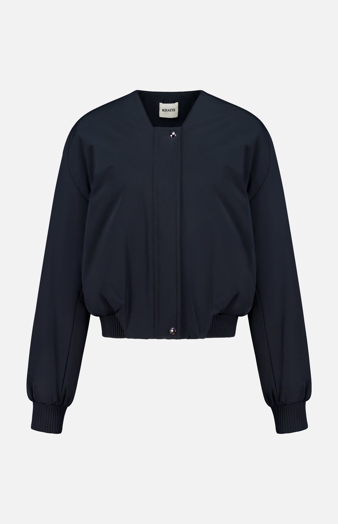 The Cordan Cropped Bomber Jacket by KHAITE is showcased against a plain white background, featuring a navy blue hue, long sleeves, ribbed cuffs and hem, and a front snap button closure—an ideal addition to any minimalist wardrobe.