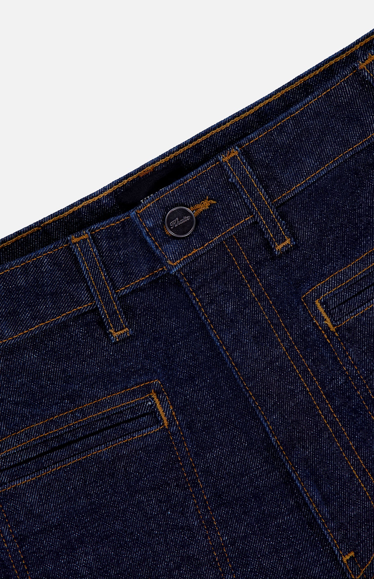 Close-up of the waistband and front pockets of the KHAITE Delmonico Wide Leg Jean in Benson, featuring high-waisted, dark wash denim with orange stitching, belt loops, and a metal button closure against a white background.