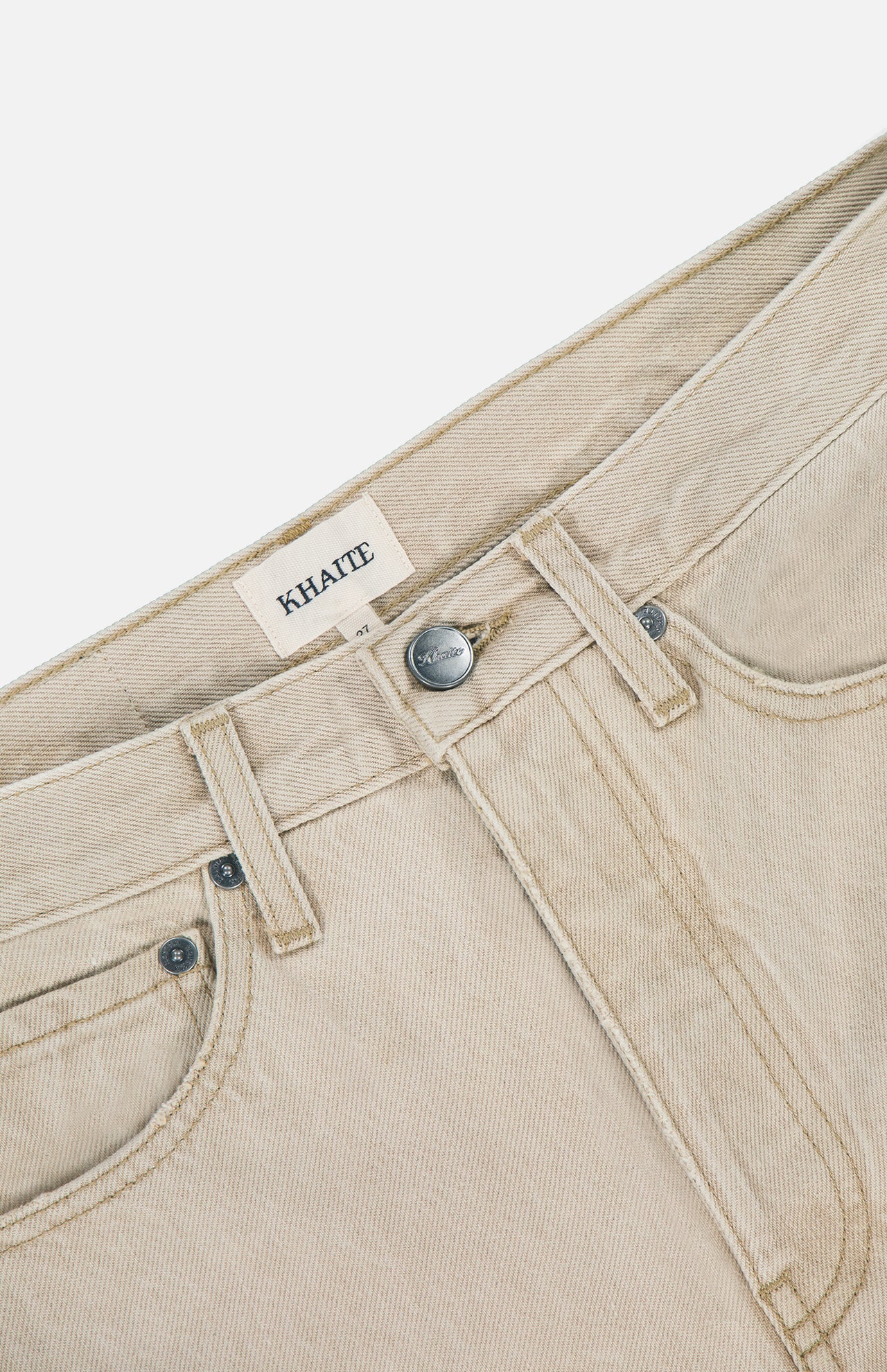Close-up of the Finley Jean in Dumas by KHAITE, featuring a mid-rise waistband, front pockets, metal button, and inner KHAITE label. Crafted from stretch-free denim with visible stitching and a lightly textured beige finish.
