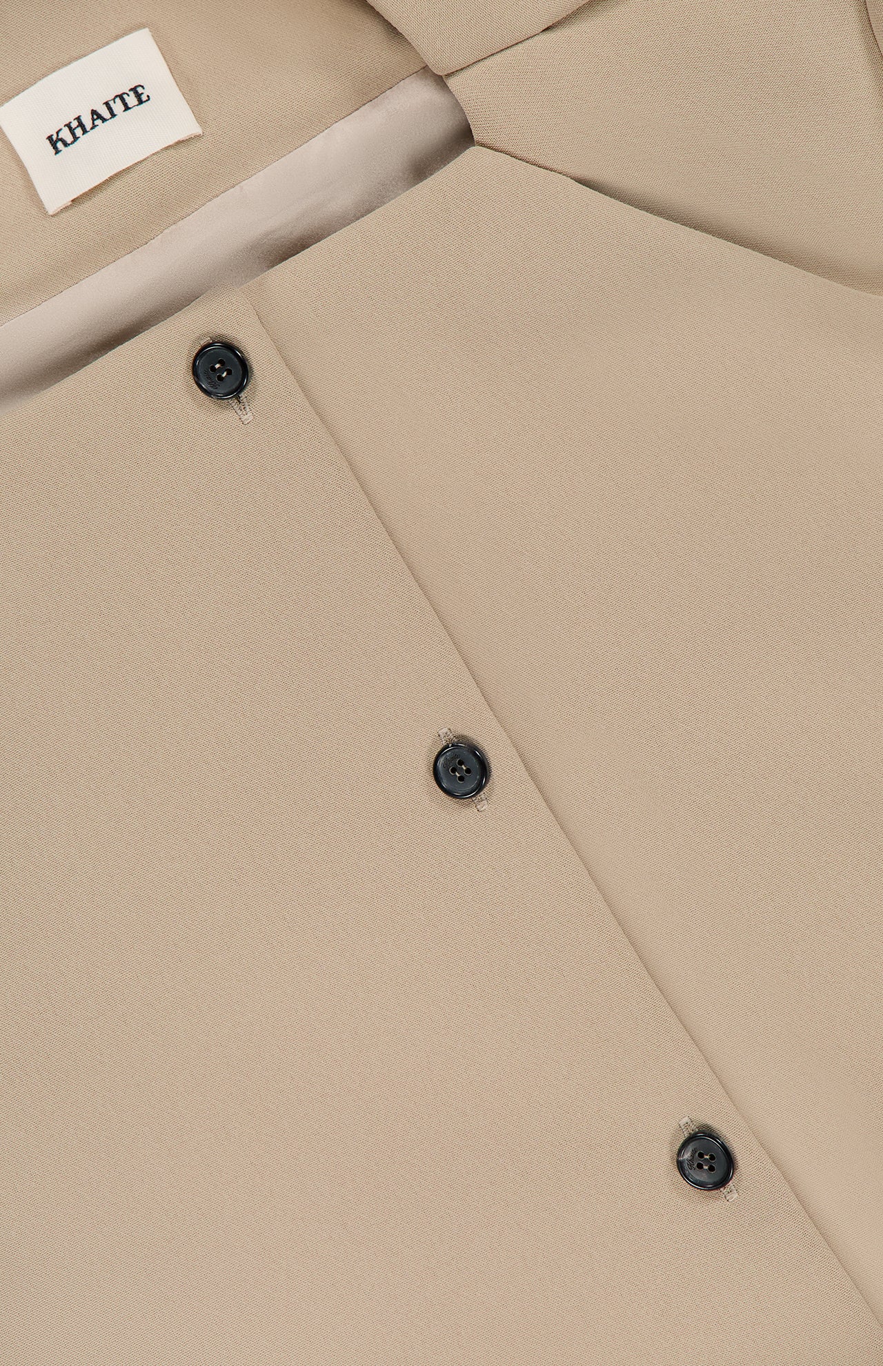 Close-up of the Folded Neck Franc Jacket by KHAITE, crafted from smooth beige Italian crepe with black buttons and a white brand label at the collar—an ideal choice for a minimalist wardrobe.