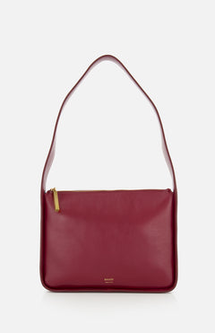 The Lori Squared Shoulder Bag by KHAITE is a minimalist rectangular burgundy leather bag with a wide strap and gold zipper, featuring a simple design and gold brand embossing near the bottom. Made in Italy, it's perfect for a minimalist wardrobe.