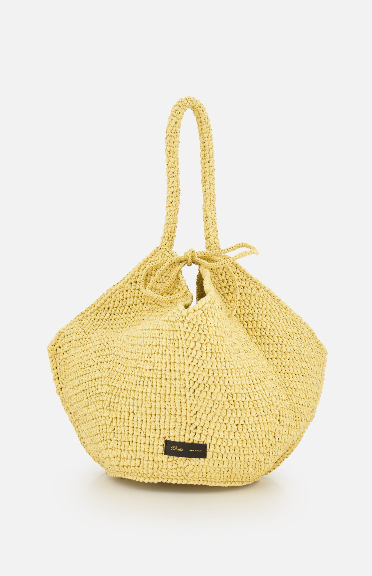 The Lotus Viscose Raffia Medium Bag by KHAITE is a yellow woven tote with two handles, a gathered drawstring top, and a small black rectangular label near the base. It’s crafted from sustainably harvested raffia.