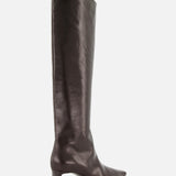 The Ona Knee High Boot by KHAITE is a tall, dark brown leather boot crafted in Italy, featuring a knee-high silhouette, pointed toe, and low block heel. The boot is displayed in side profile against a plain white background.