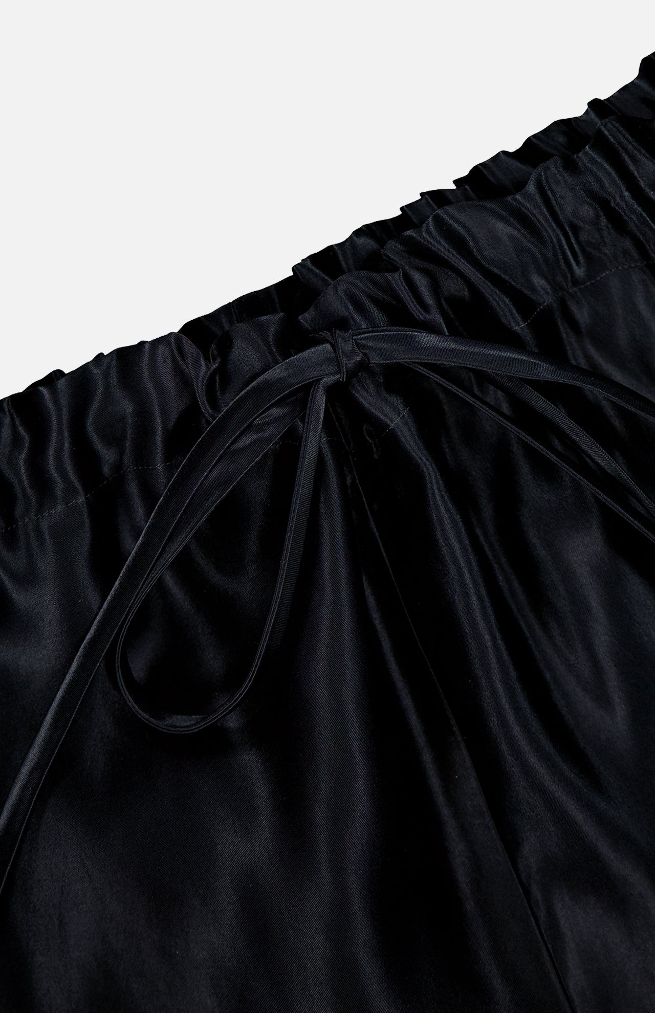 Close-up of the waistband on KHAITE’s Rohen Satin Bow-leg Pant, showcasing a gathered elastic top and central drawstring bow. The luxe black satin finish adds sophistication to any minimalist wardrobe.