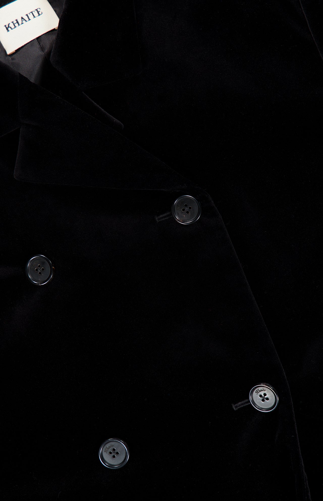 Close-up of the Rolon Velvet Coat by KHAITE—a long black cotton velvet coat featuring a double-breasted design with four black buttons and a visible KHAITE label inside the collar. A timeless addition to any minimalist wardrobe.