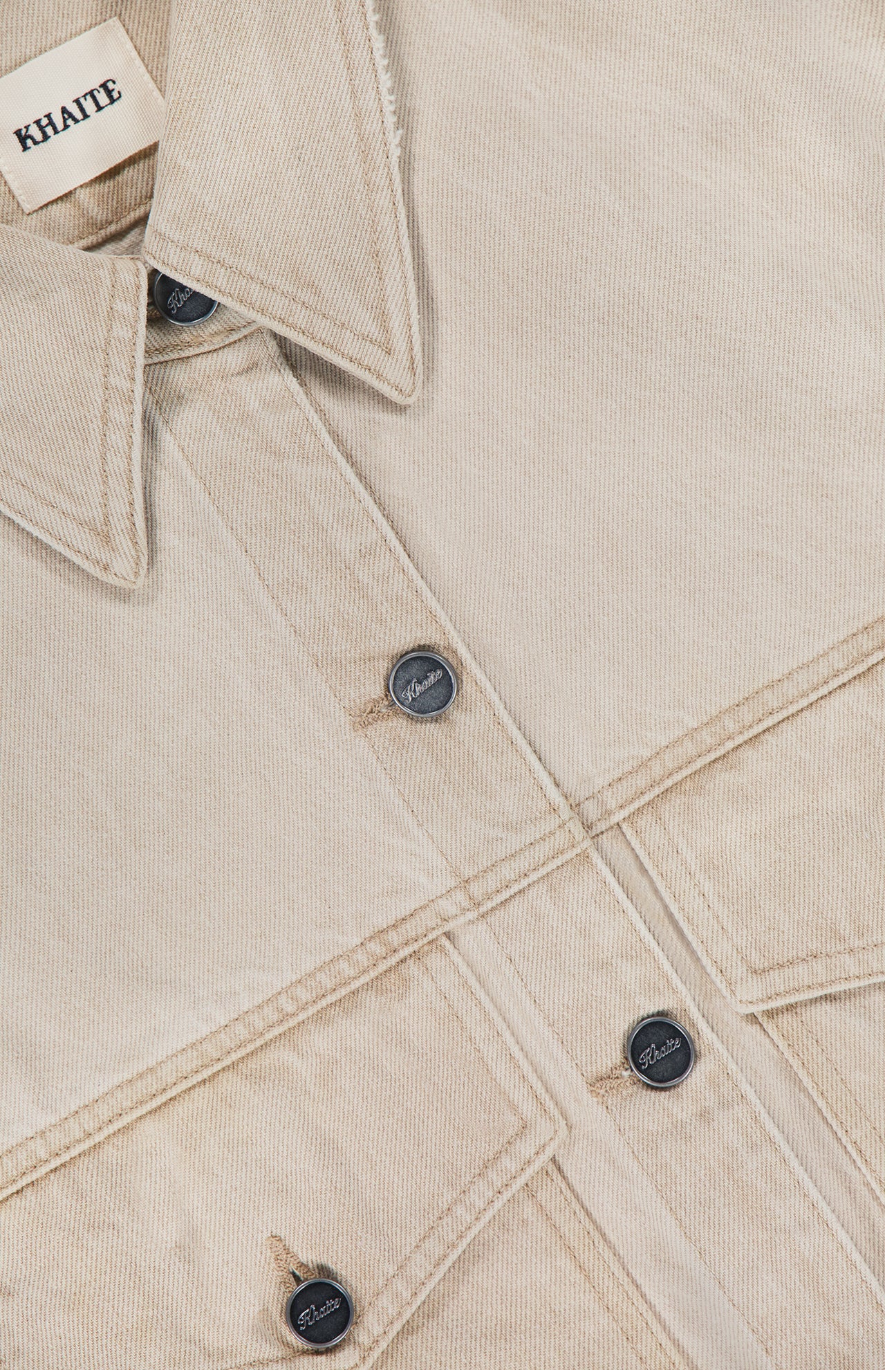 Close-up of the Tomer Collared Cropped Denim Jacket by KHAITE in beige denim, featuring black buttons and two front pockets. A white label inside the collar displays the brand name KHAITE—ideal for a minimalist wardrobe.