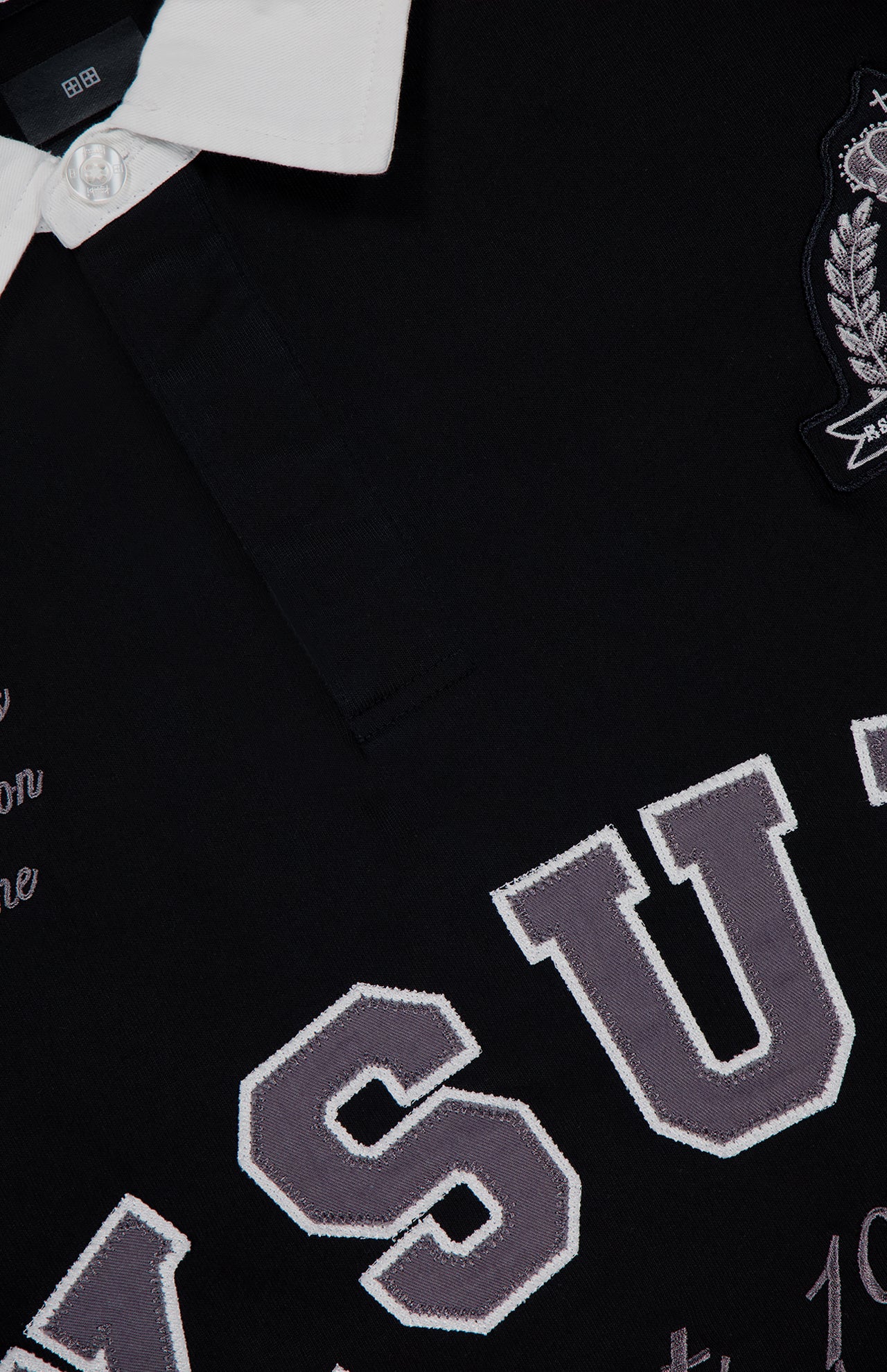 Close-up of the KSUBI Duke Rugby Long Sleeve Jersey Shirt: black with a white collar and button, featuring large gray and white embroidered collegiate-style lettering, a crest patch, and smaller decorative text on this oversized top.