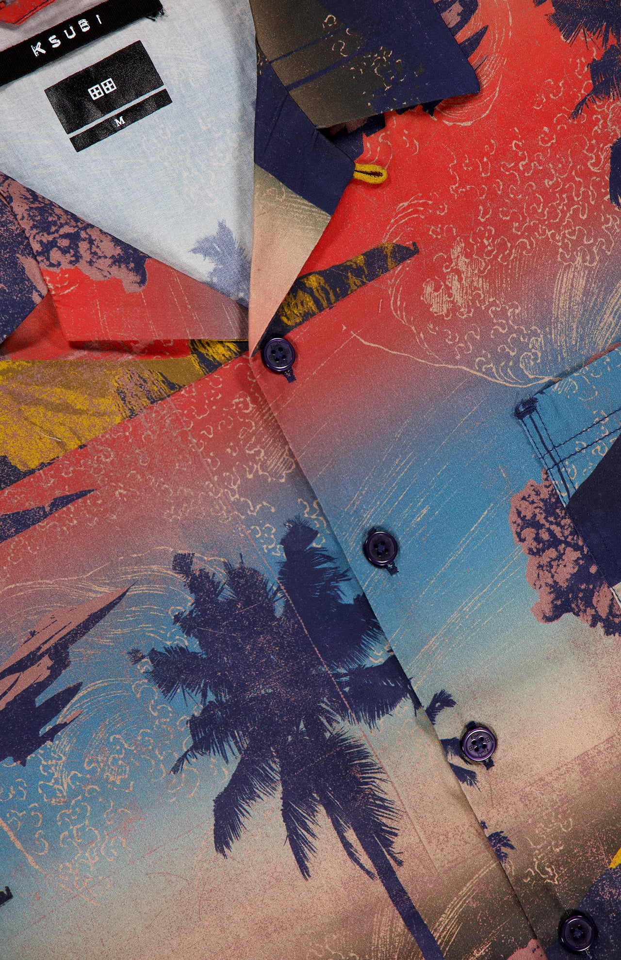 A close-up of the KSUBI Mauna Loa Short Sleeve Diner Shirt in size M, showcasing its vibrant tropical sunset print with palm trees, ocean waves, and sailboats. Made from soft cotton modal fabric; brand and size appear on the shirt tag.