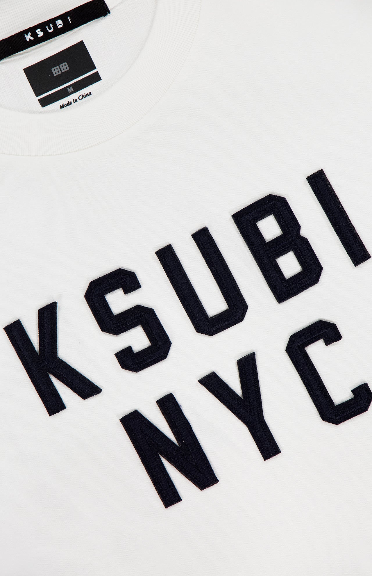 Close-up of the KSUBI NYC Ekcess Short Sleeve T-shirt in white premium cotton jersey, featuring bold black NYC logo artwork on the front and a collar tag with the KSUBI brand name.