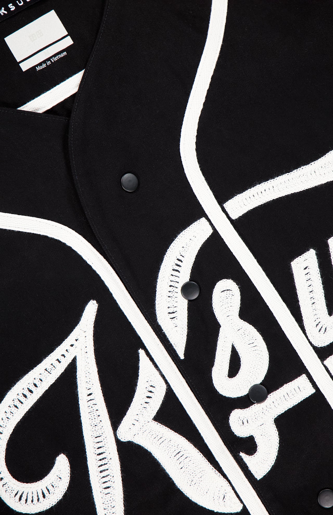 Close-up of the KSUBI Outfield Short Sleeve Jersey Top in black, featuring bold white embroidered lettering and trim. The partially visible text and shirt tag indicate it is made in Vietnam.